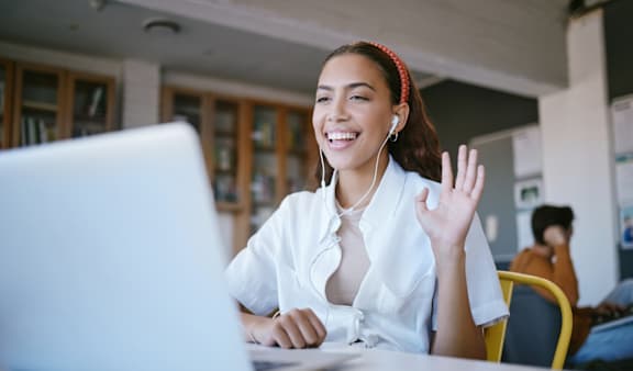 A woman in a white shirt is sitting at a table with a laptop and waving.