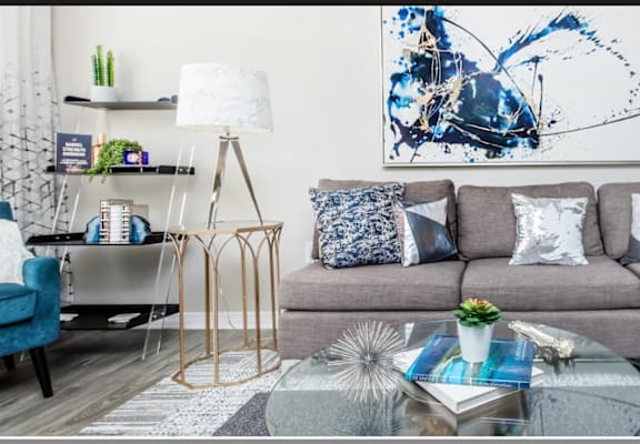 A living room with a grey couch, a blue chair, a glass table, and a large abstract painting on the wall.