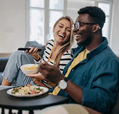 A man and a woman are sitting on a couch, eating and watching TV.
