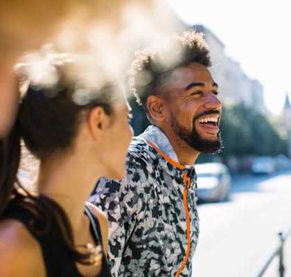 A man with a beard and a patterned shirt is laughing while standing on a street.
