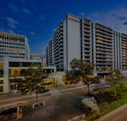 A modern building with a glass facade is surrounded by trees and a busy street with cars.