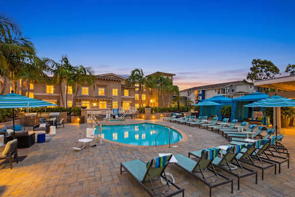 A poolside area with lounge chairs and umbrellas is illuminated at dusk.