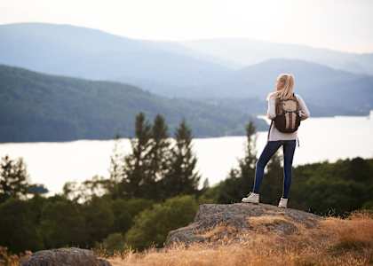 A woman with a backpack stands on a rock overlooking a lake and mountains.