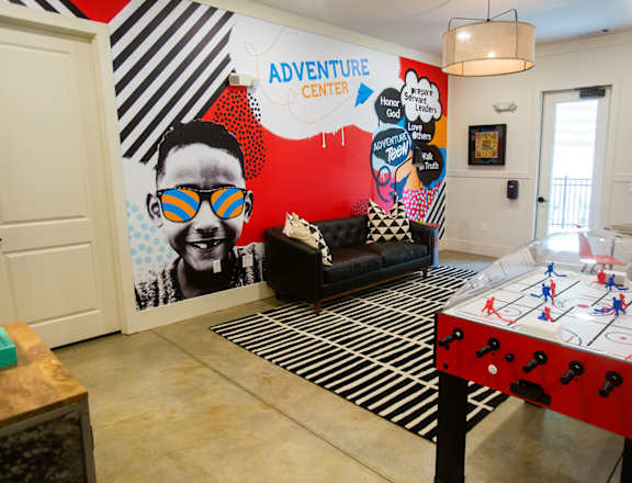 A room with a red and white air hockey table in front of a wall with a mural of a man wearing sunglasses.