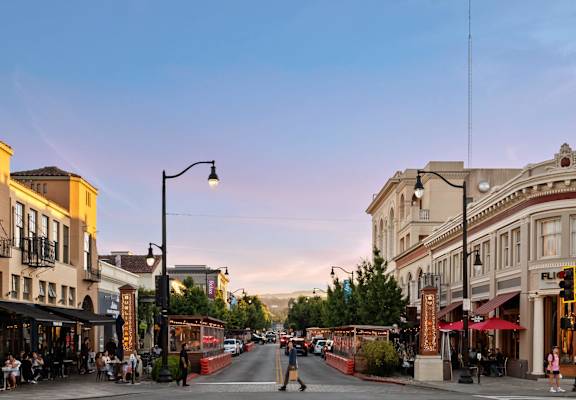 A street view of a city with buildings on either side and people sitting at tables outside the restaurants.