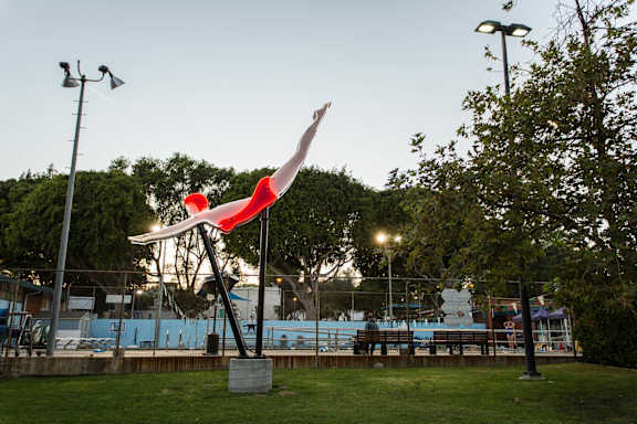 A red and white sculpture of a bird is in the foreground of a park with a pool in the background.