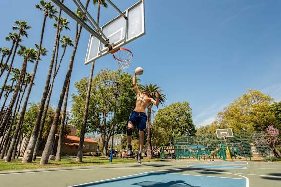 A man is playing basketball on a court with palm trees in the background.
