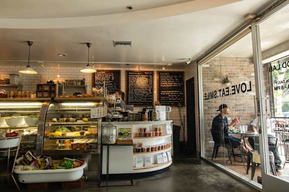 A man is sitting at a table in a cafe with a display case full of food behind him.