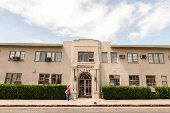 A person stands in front of a beige building with a green roof.