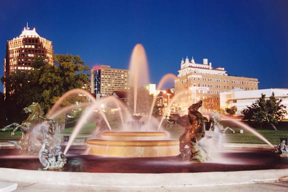 A fountain in the middle of a park with buildings in the background.