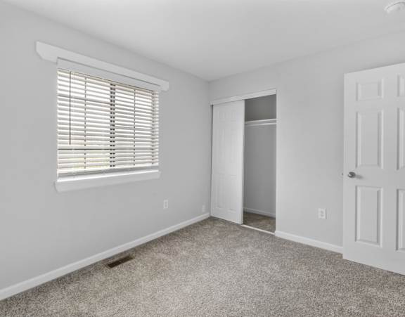 A room with a white door, a window with blinds, and a carpeted floor.