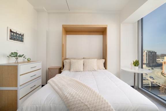 A bedroom with a large bed, a nightstand, and a window overlooking a cityscape.