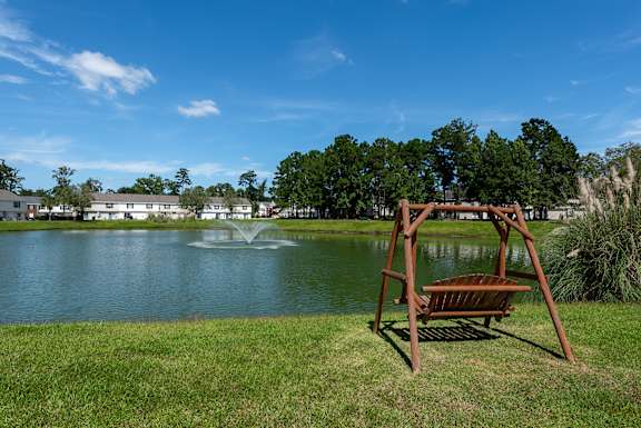 A wooden bench is in the foreground of a park with a pond and a fountain.