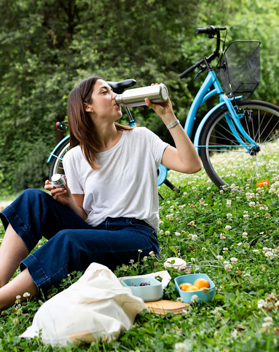 A woman is sitting on the grass and drinking from a metal bottle.