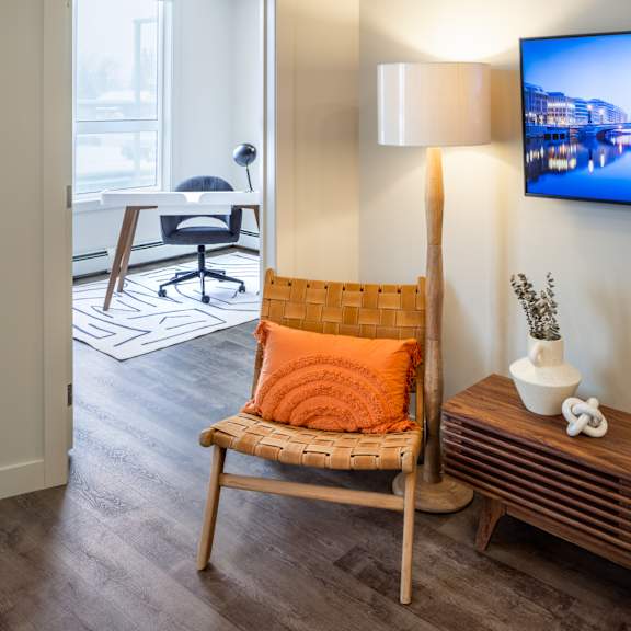 A chair with an orange pillow is in the foreground of a room with a television on the wall.