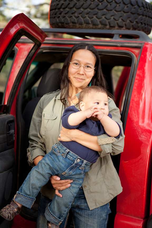 A woman holding a baby in her arms is standing in front of a red car.