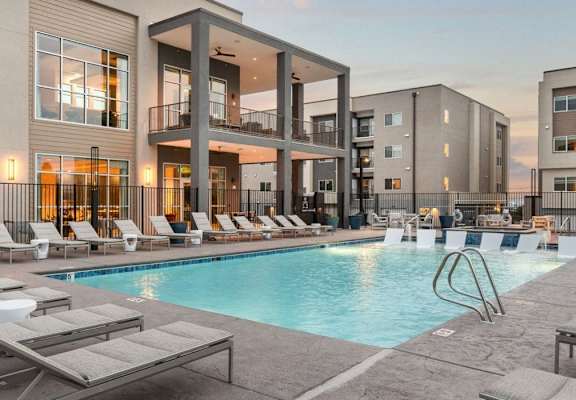 A swimming pool surrounded by sun loungers in front of apartment buildings.
