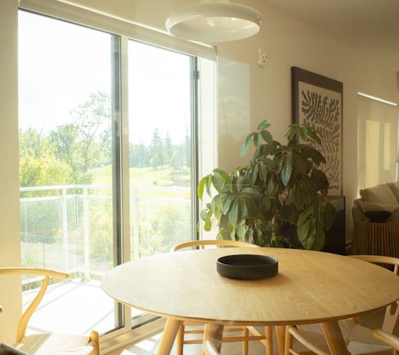 A round wooden table with a bowl on it is in the center of a room with a large window.