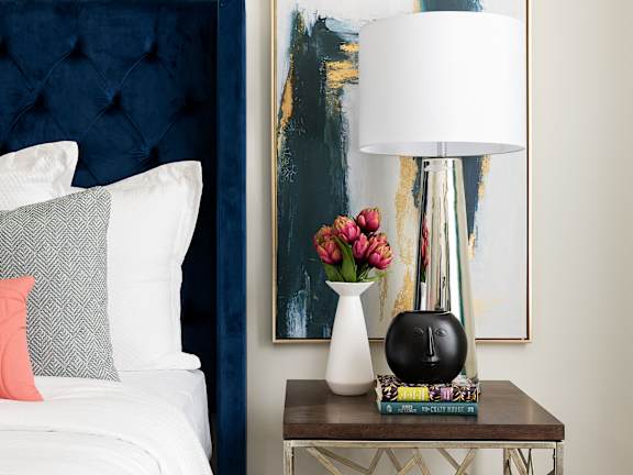 A bedroom with a bed, a side table with a lamp and a painting on the wall.