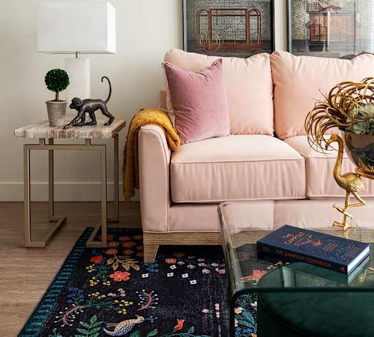 A living room with a pink couch, a small table with a plant and a cat figurine, and a rug with a colorful pattern.