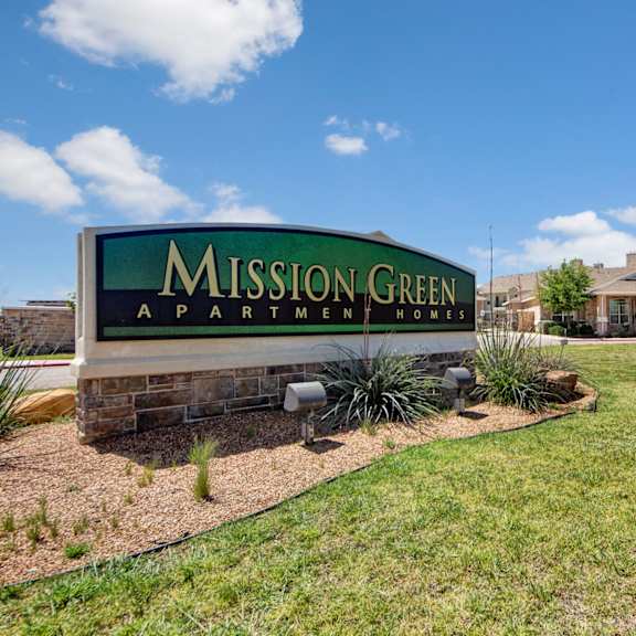 The entrance sign to Mission Green apartments surrounded by green grass and some plants. 