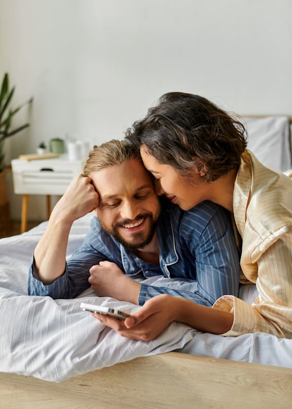 A man and woman are lying on a bed looking at a smartphone.