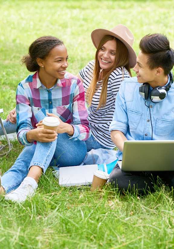 Three people sitting on grass with a laptop and headphones.