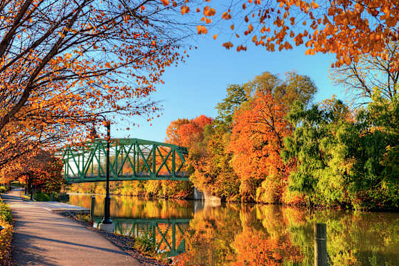 A pathway with a bridge over a lake surrounded by trees with orange leaves.