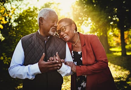An older couple standing in a park with the man wearing a brown vest and the woman wearing a red blazer.