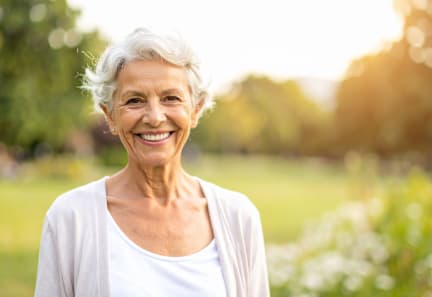 An elderly woman with short white hair is smiling in a park.