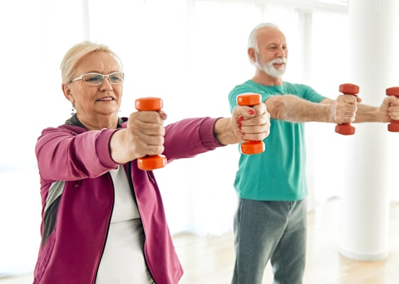 Two elderly people exercising with dumbbells in a bright room.