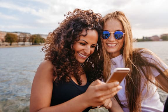 Two women are looking at a smartphone together by the water.