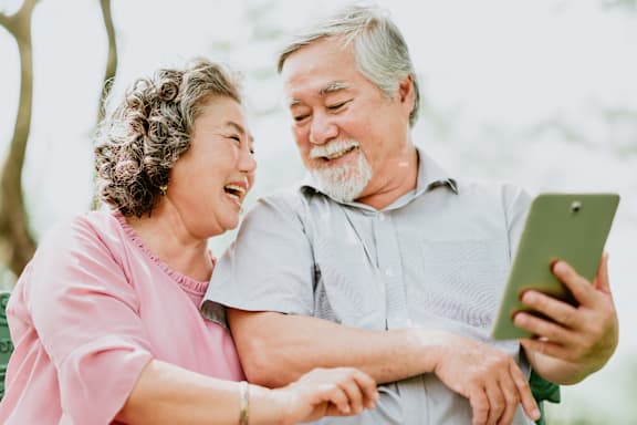 An elderly couple is sitting on a bench with a tablet in the man's hand.