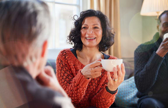 A woman in a red sweater is holding a white cup and smiling while two other people sit across from her.