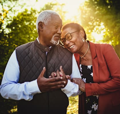 An older couple standing in a park with the man wearing a brown vest and the woman wearing a red blazer.