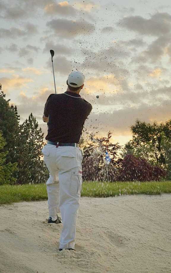 A man in a white cap and dark shirt is playing golf on a sandy field.