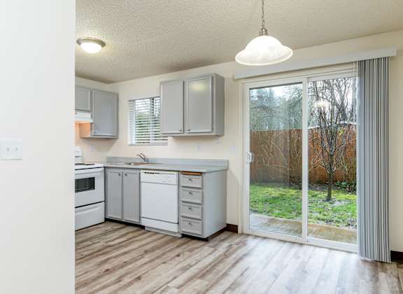 A kitchen with white appliances and cabinets with a sliding glass door leading to a backyard.