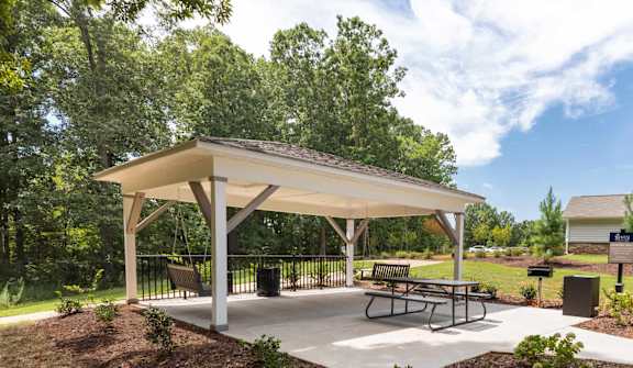 A pavilion with a white roof and a picnic table is surrounded by a mulched garden bed.