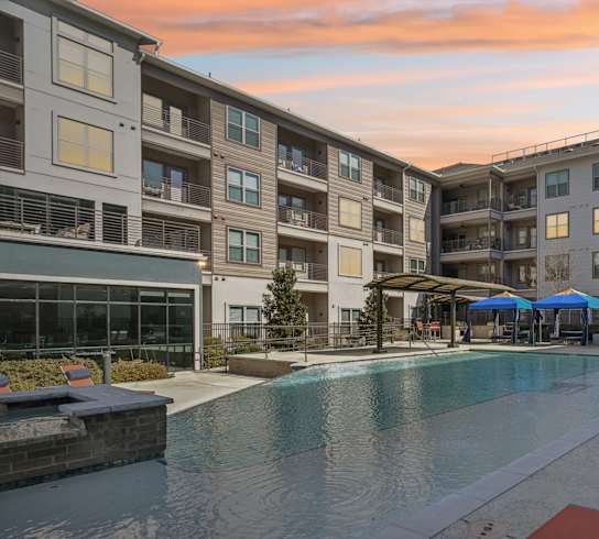 A swimming pool surrounded by sun loungers and umbrellas in front of apartment buildings.