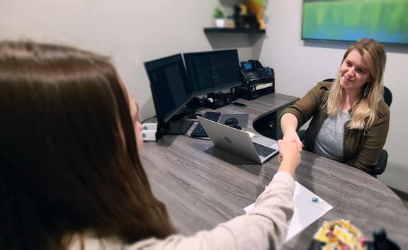 a woman sitting at a desk with a laptop and another woman talking to a colleague