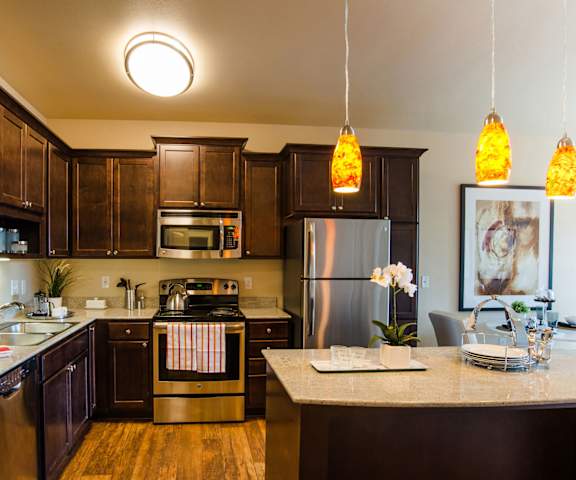 A large, modern kitchen with stainless steel appliances, island with barstools, decorative orchid, kitchen utensils, marble countertops with lots of room, unique lighting, and large wooden cabinets at Siena Apartment Homes. 