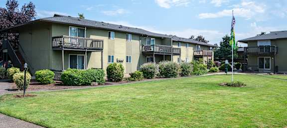A grassy area in front of apartment buildings with bushes and a flag.