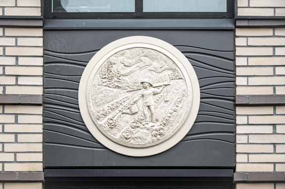 A circular relief sculpture of a man holding a spear is mounted on a brick wall.