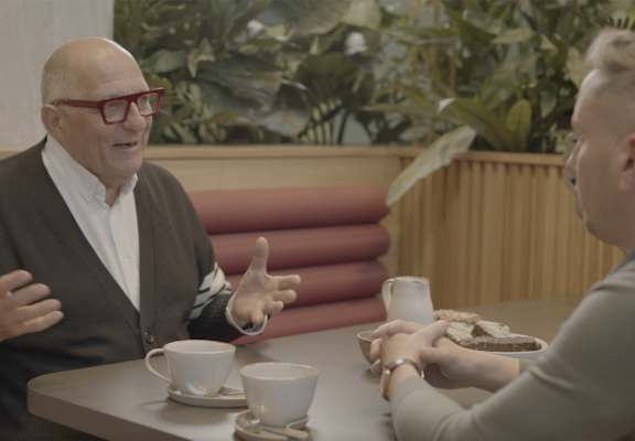 A man in a brown jacket and red glasses is gesturing with his hands while sitting at a table with two cups of coffee and a sandwich.