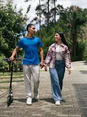 A man and a woman are walking together, the man is holding a scooter.