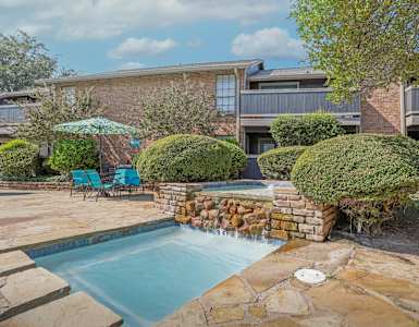 A pool surrounded by stone and bushes with a building in the background.
