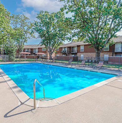 A partly shaded swimming pool surrounded by trees and chairs with umbrellas at Orleans Square in Shreveport, LA