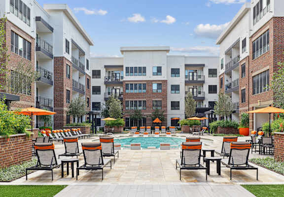 A pool surrounded by chairs and umbrellas in front of apartment buildings.