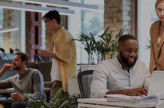 A group of people in a modern office setting with one person working on a laptop and others standing around a table.