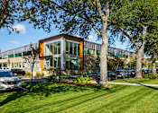 Thumbnail 12 of 21 - A modern building with orange and green accents is surrounded by trees and parked cars.